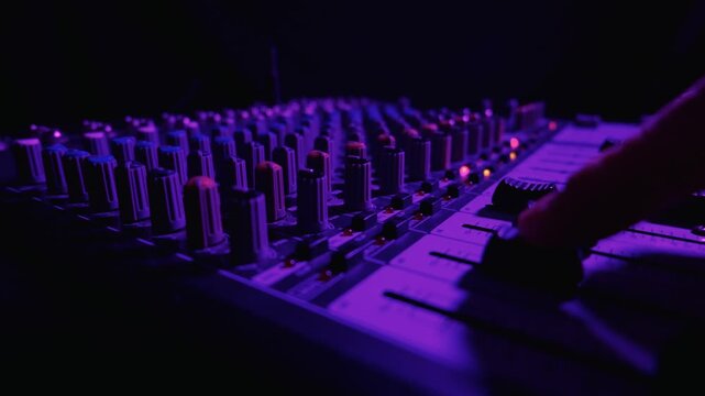 A professional audio mixing console shines under deep purple studio lights. Rows of knobs and volume faders extend into a dark, softly blurred background. Red indicator lights glow.
