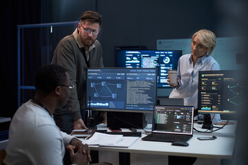 Multiethnic business team reviewing cybersecurity data on monitors, discussing network analysis in office. Ideal for IT operations, threat detection, digital security marketing © DragonImages