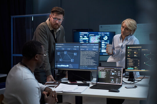 Multiethnic business team reviewing cybersecurity data on monitors, discussing network analysis in office. Ideal for IT operations, threat detection, digital security marketing