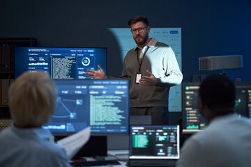 Obraz premium Medium shot of Caucasian young adult man presenting cybersecurity data to team, explaining code and network monitoring workflow. Ideal for IT training, SOC operations, tech business
