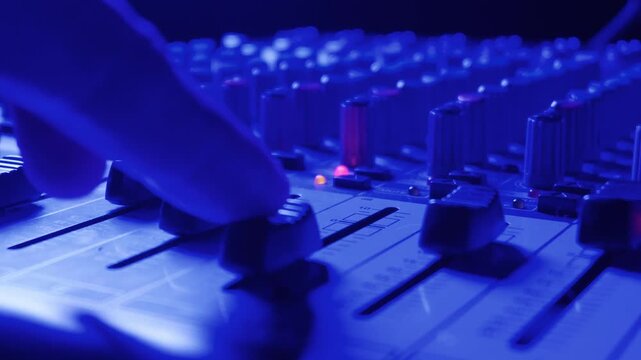 Hand moves a black fader on an audio mixing desk. Scene is illuminated with blue light and shows several knobs in the background. Close-up view highlights the sound equipment used.