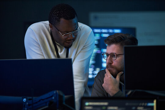 African American man and Caucasian man reviewing code on desktop monitors, collaborating on problem solving in IT office. Ideal for cybersecurity, software development, teamwork marketing