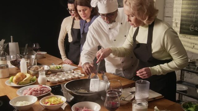 High angle shot of middle aged woman stirring onions in pan while chef helping her during group cooking master class in restaurant