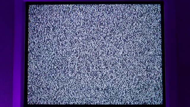 Analog static noise displays on a vintage CRT television screen illuminated by purple ambient light. Concept of signal interference and retro media technology in a dark setting.