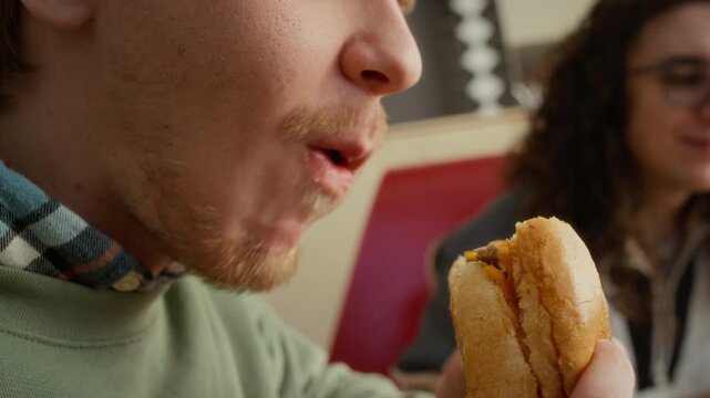 Extreme close-up of lower face of young Caucasian zoomer guy biting off mouthfuls of delicious cheeseburger, chewing with delight and pleasure while indulging in fast food at diner