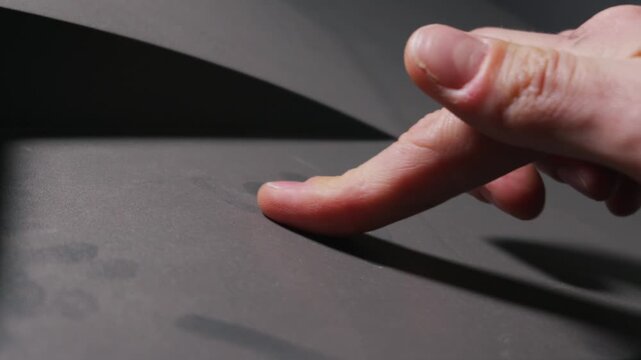 A human finger gently touches a dark grey matte textured surface. Diffused studio lighting highlights subtle grain and intricate tactile interaction. Delicate skin contact is visible,