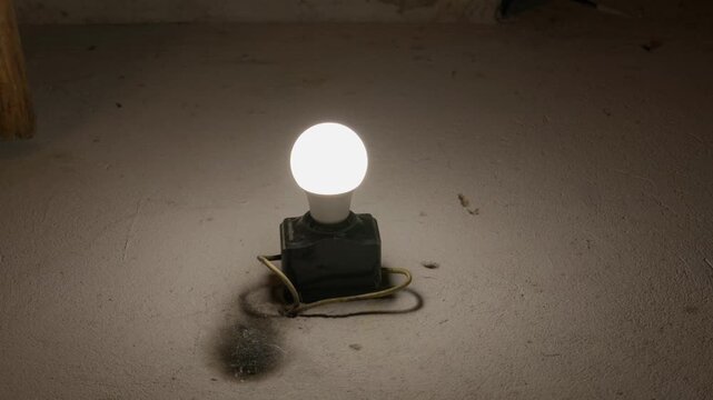 A glowing LED light bulb shines brightly from an old, dirty black socket. Exposed wiring wraps around the base on a rough, grunge textured wall. A dark wooden beam is visible.
