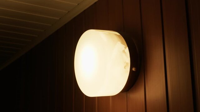 A warm glowing round light fixture shines brightly on a dark vertically paneled exterior house wall at night. Concept of evening ambiance, home security, and warm outdoor illumination.