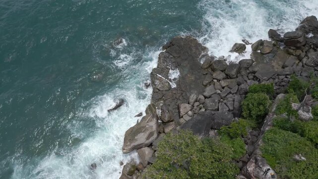 Ocean waves strike a rocky outcrop below steep cliffs. Seen from above in a rotational camera movment.