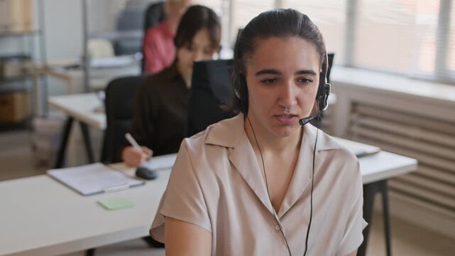Handheld shot of young Caucasian support agent in headset providing help while sitting at desk in call center with colleagues in background