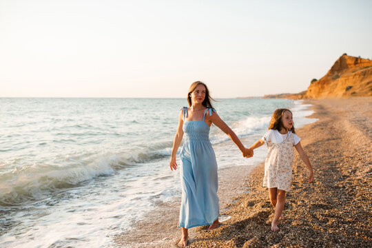 Happy woman with child girl daughter 5-6 year old holding hands walking at beach over sea at background. Summer vacation time. Motherhood.