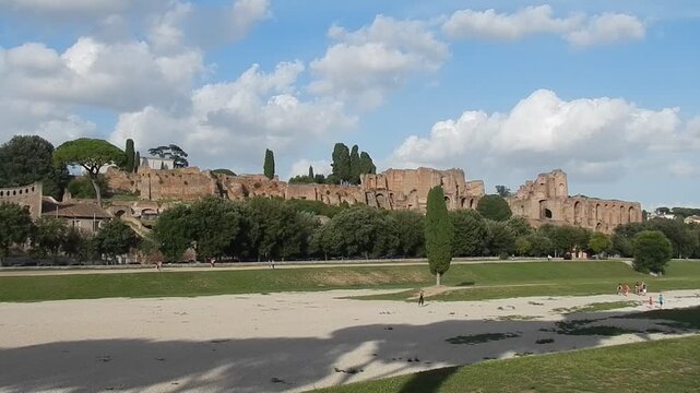 Ancient Rome in Italy - view with Palatine Hill and Circus Maximus, green trees of Mediterranean vegetation, blue sky with white clouds and silhouettes of people passing on August 2014.