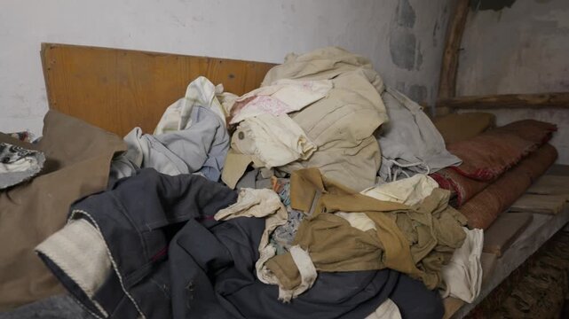 Various worn garments and patterned fabrics lie in a cluttered pile on a wooden shelf against a rustic wall. Concept of poverty, disorganized storage, and abandoned domestic life.