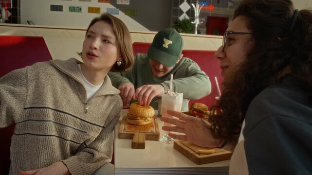 Medium shot of young male zoomers playing practical joke on female friend, distracting by conversation, adding spicy sauce to burger then laughing while girl drinking milkshake to reduce burning