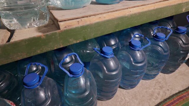 A close-up view of numerous large plastic water bottles with blue caps stored in a row under a green wooden shelf. Sunlight highlights the clear water containers on the ground.