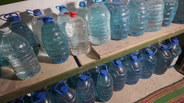Numerous large plastic bottles filled with drinking water rest on wooden storage shelves. Concept of emergency preparedness, essential survival supplies, and resource hoarding.