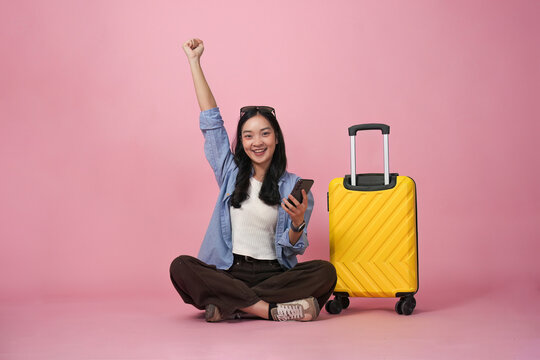 Asian woman celebrating travel booking success with smartphone and luggage