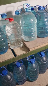 Large plastic jugs filled with clear drinking water sit stored on wooden shelves in a dim pantry room. Concept of emergency preparedness, survival supplies, and basic necessities.