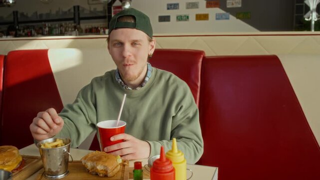 Medium slowmo portrait of young Caucasian male student, wearing baseball cap, chewing fries and drinking soda while enjoying fast food meal with cheeseburger at favorite diner, looking at camera
