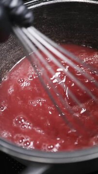 Chef batiendo l&iacute;quido rojo infusionado en una olla negra de hierro fundido. Chef profesional revolviendo la infusi&oacute;n roja con un batidor de metal. Olla de hierro fundido, proceso culinario artesanal.