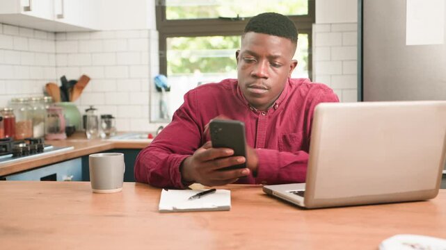 Focused African American Man Working Remotely From Home Kitchen with Laptop and Smartphone