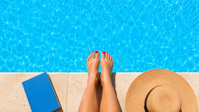 Bare feet resting on a swimming pool edge with a book and straw hat