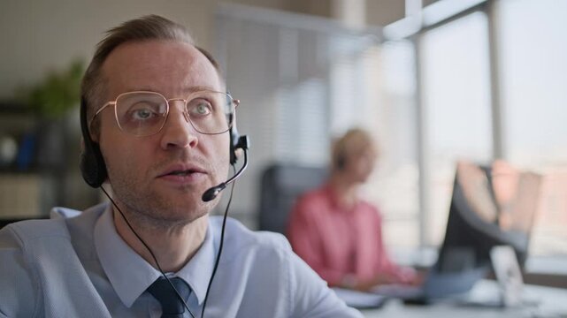 Chest up stab shot of stress-resistant Caucasian support agent giving instruction to client via headset and taking off glasses while working in modern call center