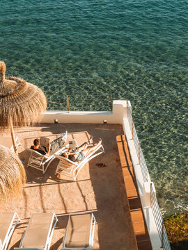View of people relaxing on sun loungers on a terrace with straw umbrellas overlooking the clear turquoise sea in Sant Elm, Balearic Islands, Spain.