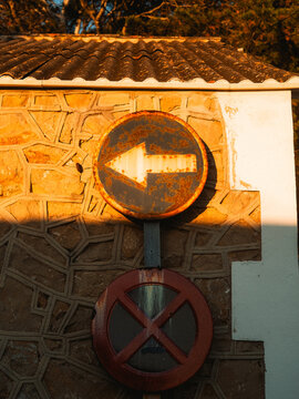 View of rusty road signs with a white arrow and a red cross on a stone wall under golden sunlight Sant Elm, Balearic Islands, Spain.