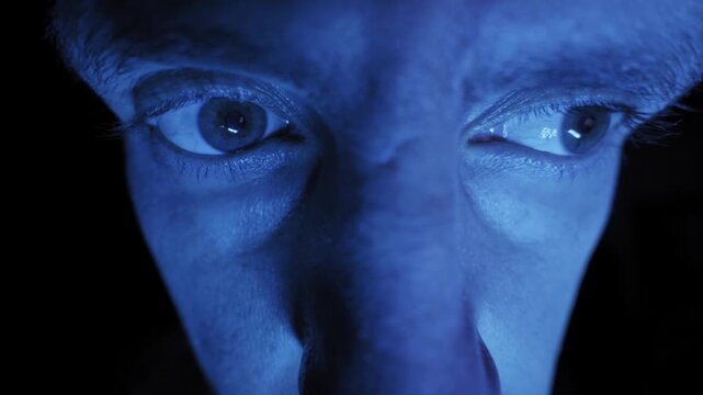 Extreme close-up of a man's eyes darting around, illuminated by a blue screen in darkness. Concept of cybersecurity, internet addiction, and digital surveillance.
