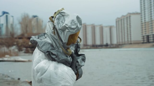 Protective suit on icy riverbank survivor searching shelter with makeshift pack desolate promenade, distant towers, muffled breath, slow steps, hopeful glance toward skyline, melancholic mood