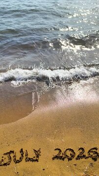 Vertical July 2026 text written on beach sand in slow motion on a Mediterranean beach at golden hour with warm cinematic light.