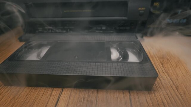 A classic black plastic VHS cassette tape sits on a wooden table near a VCR player. Dense white smoke gracefully swirls around the retro media, creating a mysterious atmosphere as it slowly