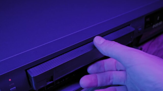 A male hand ejects a VHS tape from a vintage VCR player. The scene is illuminated by stylized violet neon lighting, evoking a retro eighties synthwave atmosphere, perfect for media and