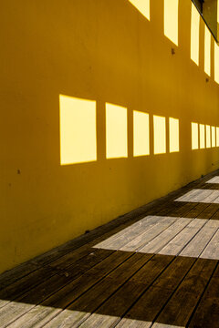 Yellow wall beside a wooden deck walkway catches square windows shadows, stretching light into architecture geometry and deep perspective