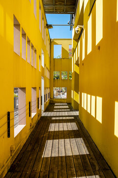Yellow courtyard walkway runs through a narrow building passage as windows cast shadows and architecture geometry repeats in hard afternoon light