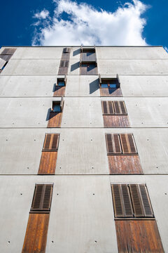 Vertical concrete building facade rises toward blue sky as wooden shutters punctuate windows in a strict architecture pattern beneath a bright cloud