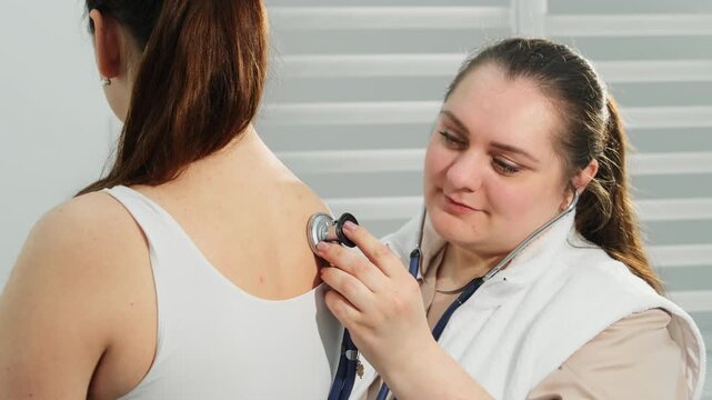 Healthcare worker performs auscultation on upper woman back during routine exam. Nurse moves stethoscope across shoulder area listening to body sounds