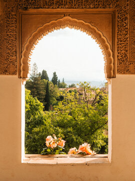 View of the Alhambra gardens through an ornate Moorish arch with intricate carvings and blooming flowers on the windowsill in Granada, Andalusia, Spain.