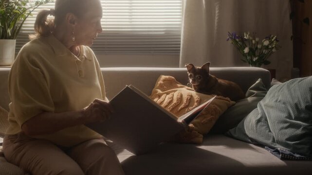 Medium shot of older Caucasian woman browsing large photograph album while sitting beside small Chihuahua dog in warm apartment interior