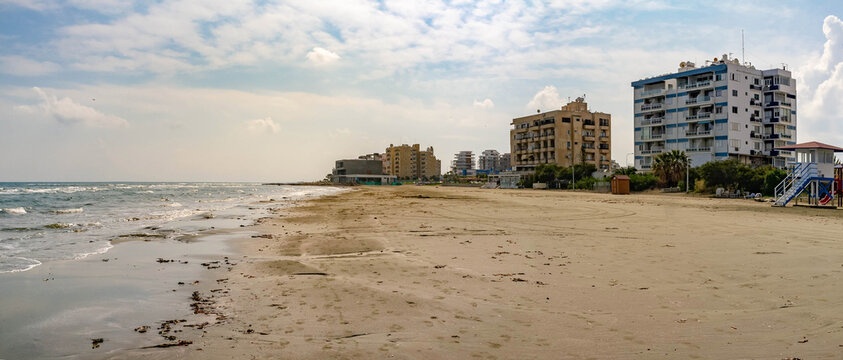 Wide panoramic view of empty sandy city beach and coastal apartment buildings in Larnaca during off-season. Sea waves on shore under cloudy sky. Cyprus.
