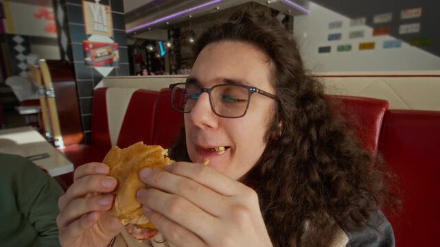 Zoom-in closeup of face of young Caucasian male zoomer with curly brown hair and eyeglasses munching on delicious hamburger with full mouth while eating out with friends at retro American diner