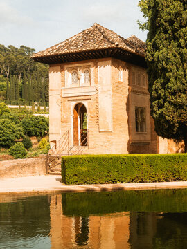 View of the historic Alhambra palace pavilion reflected in a calm pool surrounded by lush gardens and cypress trees in Granada, Andalusia, Spain.