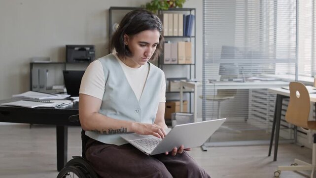 Medium long portrait of cheerful Caucasian manager in wheelchair typing on laptop and looking at camera with smile while working in office