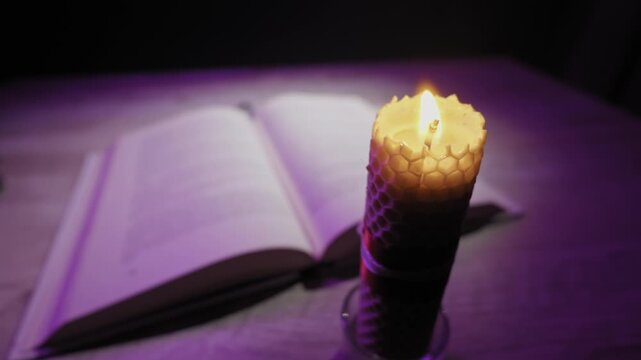 A burning beeswax candle with a honeycomb pattern flickers in front of an open book. The dark scene is lit by the flame and a mysterious purple light, creating a spiritual or magical mood.