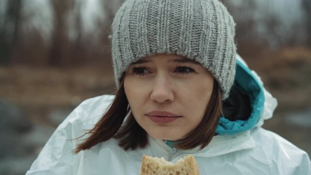 White woman survivor holding ration outdoors on icy shore, knit hat and white jacket, tired steady gaze toward distant treeline, windtossed hair, quiet melancholy and hopeful endurance, muted color