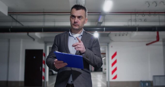 Man inspector compares completed work in underground parking with written data recorded on page. Businessman in formal attire reviews structured report attentively