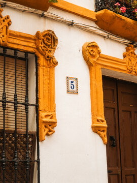 View of ornate yellow window frames, a wooden door and pink flowers on a traditional white building facade in Cordoba, Andalusia, Spain.