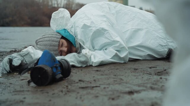 Child lying in hazmat suit shore, gasmask beside, riverbank bleak, rescuers in white coveralls approaching, tense cold atmosphere of nuclear winter, abandoned buildings on horizon, muddy ground