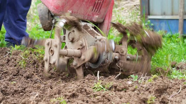 Person using rotary cultivator machine to till and churn up soil in garden area, preparing earth for future planting through manual agricultural work in springtime countryside.